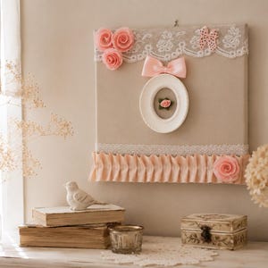Décoration murale artisanale de style shabby chic composée d'une toile beige ornée de dentelle ancienne et d'un ruban rose chinés sur un marché français, de fleurs en tissu, d'un papillon décoratif et d'un médaillon blanc avec une rose miniature.
