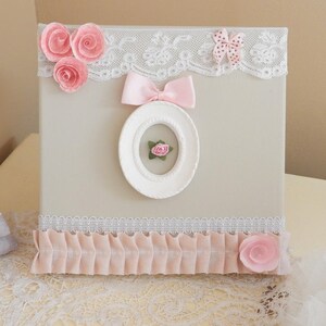 May include: A decorative square box with a light beige surface, adorned with pink fabric roses, a pink bow, and a white oval frame. The box features white lace trim and a pink ruffled border, creating a romantic, feminine aesthetic.