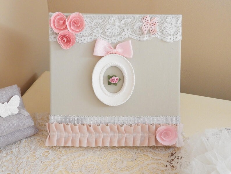 May include: A decorative square box with a light beige surface, adorned with pink fabric roses, a pink bow, and a white oval frame. The box features white lace trim and a pink ruffled border, creating a romantic, feminine aesthetic.