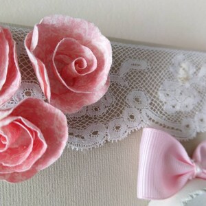May include: Close-up of a decorative piece featuring three pink paper roses with white accents. A white lace trim and a small pink bow are also visible, creating a delicate and feminine aesthetic.