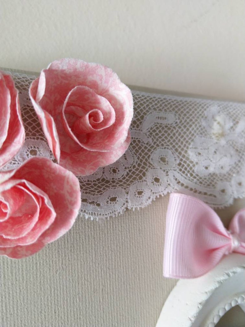 May include: Close-up of a decorative piece featuring three pink paper roses with white accents. A white lace trim and a small pink bow are also visible, creating a delicate and feminine aesthetic.