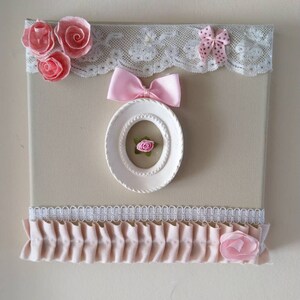 May include: A decorative canvas with a beige background, adorned with pink fabric roses, bows, and a white oval frame. The top edge features white lace trim. A ruffled pink fabric strip and a pink fabric rose are at the bottom.