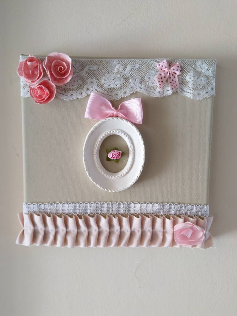 May include: A decorative canvas with a beige background, adorned with pink fabric roses, bows, and a white oval frame. The top edge features white lace trim. A ruffled pink fabric strip and a pink fabric rose are at the bottom.