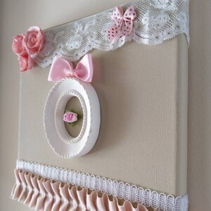 May include: A decorative canvas art piece with a beige background, adorned with pink fabric roses, bows, and lace trim. A white circular frame with a small pink rose is centered on the canvas. The piece has a romantic, feminine aesthetic.