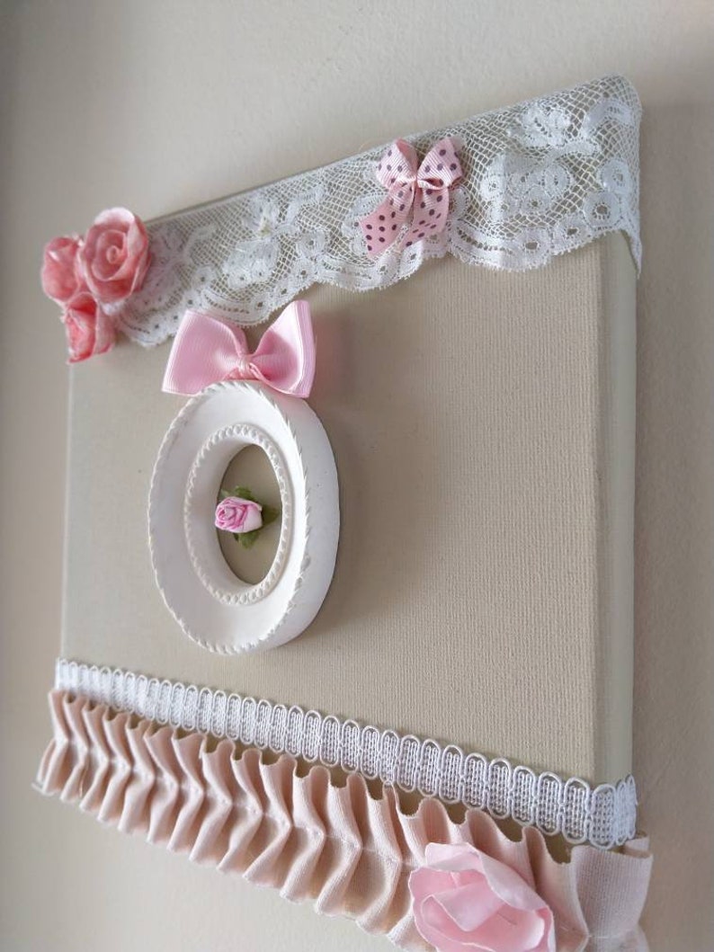 May include: A decorative canvas art piece with a beige background, adorned with pink fabric roses, bows, and lace trim. A white circular frame with a small pink rose is centered on the canvas. The piece has a romantic, feminine aesthetic.