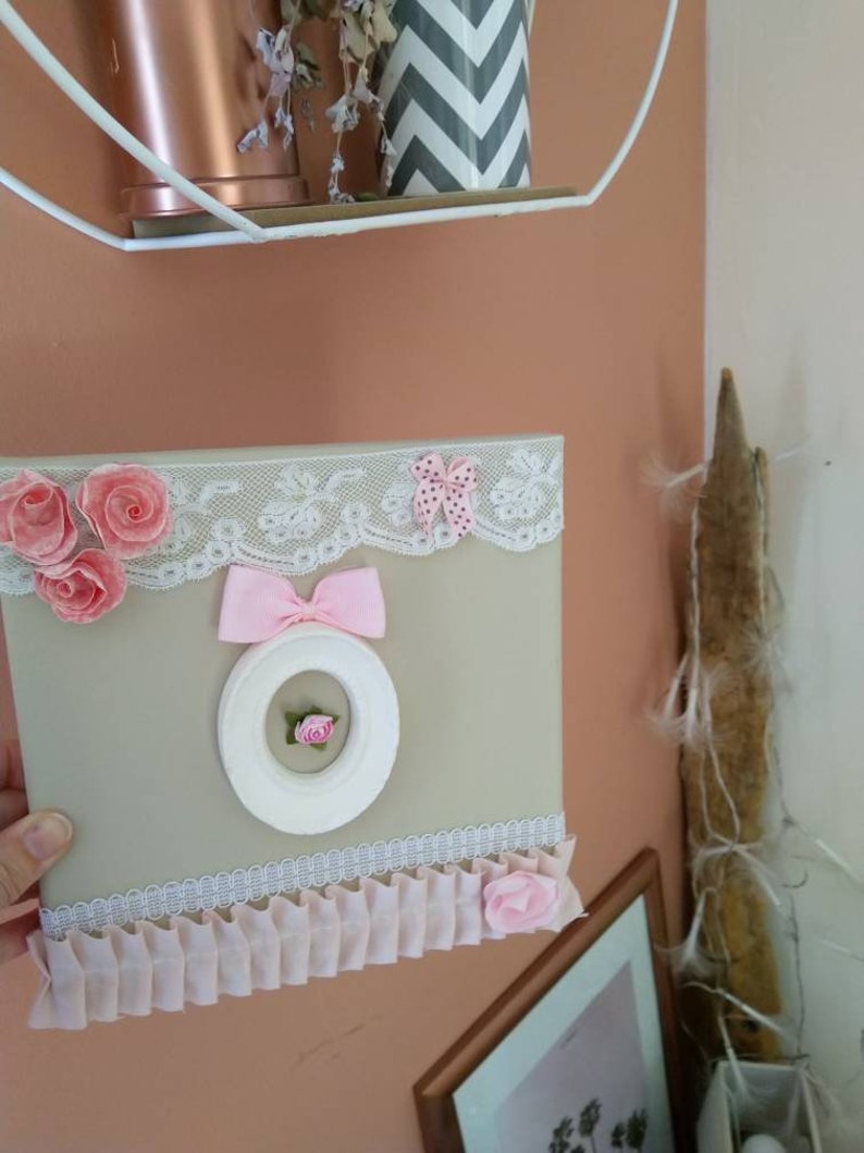 May include: A decorative framed artwork with a beige background, adorned with pink fabric roses, lace trim, and a pink bow. A white circular frame with a pink rose in the center is also featured.