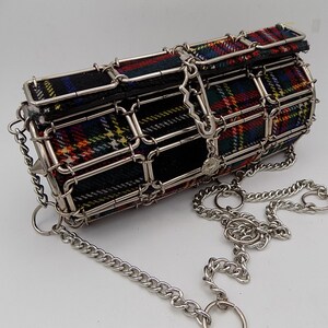 May include: A tartan patterned cylindrical handbag in red, green, yellow, and blue. The bag is encased in a silver metal cage with a chain strap and a decorative charm. The ends of the bag have silver spikes.