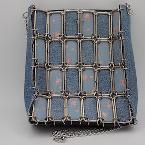 May include: A denim handbag with a silver chain strap. The bag has a grid-like metal overlay and panels of light blue fabric with a floral pattern. The bag's design combines denim with decorative metalwork.