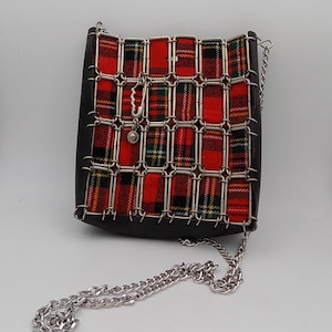 May include: A black and red plaid crossbody bag with a silver chain strap. The bag features a red plaid fabric centre, framed by a silver metal grid and black leather. A silver charm and safety pin accent the front.