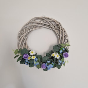 May include: A round, gray woven wreath adorned with a garland of green leaves, white daisies, blue forget-me-nots, and purple flowers. The wreath is hanging on a white wall, creating a natural and decorative accent.
