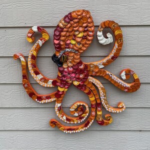 May include: A colorful octopus made from bottle caps. The octopus is orange, red, and yellow. The octopus has eight tentacles and is mounted on a white wall.