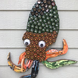 May include: A colorful octopus made from bottle caps and aluminum cans. The octopus has orange, green, and brown bottle caps for its body and tentacles. The octopus has googly eyes and is mounted on a wall.