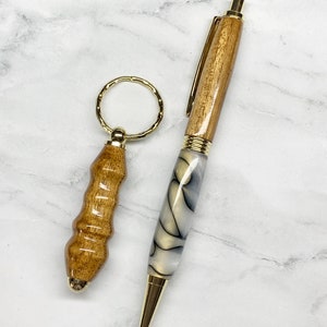 May include: A wooden pen and a matching keychain. The pen has a light wood barrel with gold accents and a marbled white and grey lower barrel. The keychain has a light wood handle and a gold keyring.