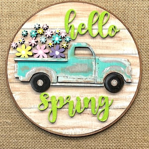 May include: A round wooden sign with a distressed teal pickup truck carrying colorful flowers. The words "hello spring" are in bright green script. The sign has a brown border and a wood-grain background.
