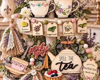 May include: A two-tiered white distressed wooden tray decorated with tea-themed items including two ceramic cups, a wooden teapot sign reading Spill the Tea, a pinky up sign, a beaded garland with tea labels, and a small fruit tart.