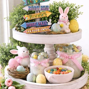 May include: A two-tiered Easter-themed display with a white wooden stand. The top tier features a signpost with directional arrows, a pink bunny figurine, and decorative eggs. The bottom tier holds jars of candy, a bowl of jelly beans, and more Easter decorations.