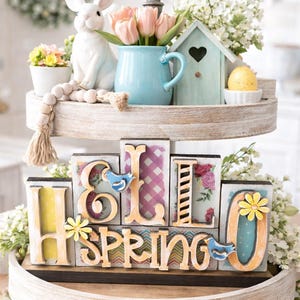 May include: A two-tiered decorative tray with spring-themed decor. The top tier holds a white bunny, a blue pitcher with pink tulips, and a small birdhouse. The bottom tier spells "Hello Spring" with decorative blocks. Greenery and eggs surround the tray.