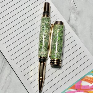 May include: A gold-toned pen with a green and white patterned barrel and cap, lying on a lined notepad. The pen's cap is off, revealing the pen tip. The notepad is decorated with colorful geometric shapes.