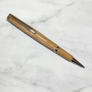 May include: A wooden pen with a light brown finish and silver-colored metal accents. The pen has a sleek design with a clip and a pointed tip. The pen is lying on a white and gray marble surface.