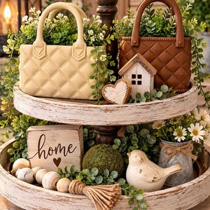 May include: A two-tiered white distressed wooden tray featuring a cream quilted handbag, a brown quilted handbag, a small wooden house, a heart cutout, a wooden block with the word home, wooden beads, a moss ball, and a ceramic bird figurine.