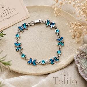May include: A silver-toned chain bracelet featuring alternating blue butterfly charms and round blue crystals. The bracelet includes a metal clasp closure. A small card with the text Telilo BIJOUX ÉNERGÉTIQUES is visible in the upper left corner.