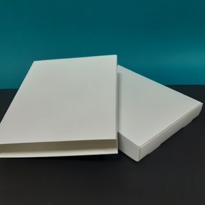 Peut inclure: Deux boîtes rectangulaires en carton blanc avec une surface lisse et plate. Une boîte est positionnée verticalement et légèrement inclinée, tandis que la seconde est posée à plat derrière, montrant un couvercle fermé et des panneaux latéraux.