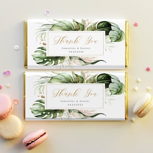 以下が含まれることがあります： 金色のアクセントが付いた白いチョコレートバーの包装が2つ。包装には、金色のアクセントが付いた熱帯の葉のデザインと、「ありがとう」の文字、サマンサ＆ダニエルの名前、2022年4月23日の日付が書かれています。