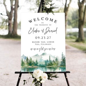 May include: A white wedding welcome sign with black text, including the names Elisha & Daniel, date 09.23.27, and location Manor Vail Lodge, Vail, Colorado. The bottom features a watercolor landscape of mountains and trees.
