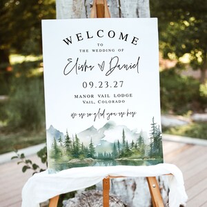 May include: A white welcome sign on an easel with the text "WELCOME TO THE WEDDING OF Elisha & Daniel 09.23.27 Manor Vail Lodge Vail, Colorado" and "we are so glad you are here". The sign has a watercolor landscape of mountains, trees, and a lake.