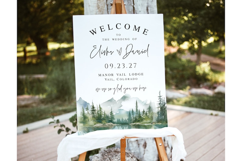 May include: A white welcome sign on an easel with the text "WELCOME TO THE WEDDING OF Elisha & Daniel 09.23.27 Manor Vail Lodge Vail, Colorado" and "we are so glad you are here". The sign has a watercolor landscape of mountains, trees, and a lake.