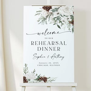 May include: A white welcome sign for a rehearsal dinner, featuring floral accents of white roses, greenery, and pinecones. The sign reads "welcome to our rehearsal dinner" with the names Sophia & Anthony and the date and location.