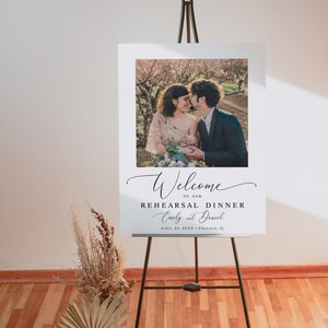 May include: White welcome sign with a photo of a couple in a garden setting. The text reads "Welcome to our Rehearsal Dinner Emily & David April 23, 2023 Chicago, IL"