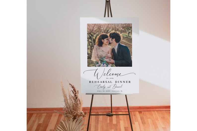 May include: White welcome sign with a photo of a couple in a garden setting. The text reads "Welcome to our Rehearsal Dinner Emily & David April 23, 2023 Chicago, IL"