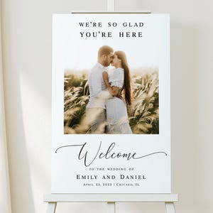 May include: A white wedding welcome sign with a black script font that reads "We're so glad you're here" and "Welcome to the wedding of Emily and Daniel". The sign also includes the date "April 23, 2022" and location "Chicago, IL". A photo of a couple kissing in a field is featured in the center of the sign.