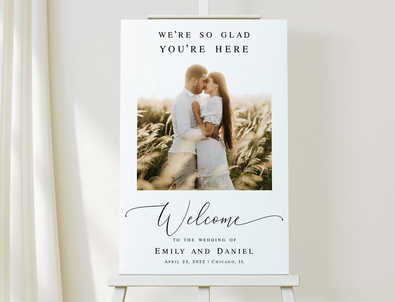 May include: A white wedding welcome sign with a black script font that reads "We're so glad you're here" and "Welcome to the wedding of Emily and Daniel". The sign also includes the date "April 23, 2022" and location "Chicago, IL". A photo of a couple kissing in a field is featured in the center of the sign.