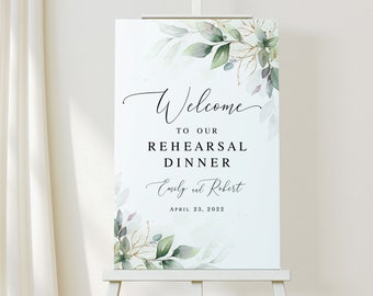 May include: A white sign with green and gold watercolor leaves and branches. The text reads "Welcome to our Rehearsal Dinner Emily and Robert April 23, 2022".