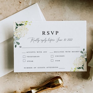 May include: A white RSVP card with a floral design featuring white hydrangeas and green leaves. The card has the text "RSVP" in black font and the text "Kindly reply before June 10, 2022" in cursive font. The card also includes a section for guests to indicate their meal preference and the number of guests attending.