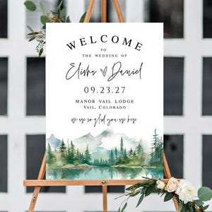 May include: A wedding welcome sign on a wooden easel. The sign features the text "WELCOME TO THE WEDDING OF Elisha & Daniel 09.23.27 Manor Vail Lodge Vail, Colorado". A watercolor landscape of mountains and trees is at the bottom.