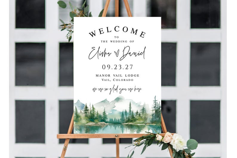 May include: A wedding welcome sign on a wooden easel. The sign features the text "WELCOME TO THE WEDDING OF Elisha & Daniel 09.23.27 Manor Vail Lodge Vail, Colorado". A watercolor landscape of mountains and trees is at the bottom.