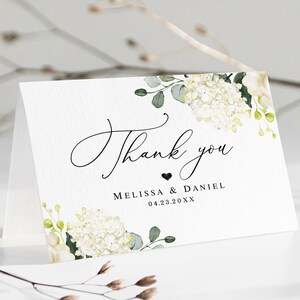 May include: White thank you card with a floral design featuring white hydrangeas and green leaves. The text "Thank you" is written in cursive with a small heart and the names "Melissa & Daniel" and the date "04.23.20XX" below.