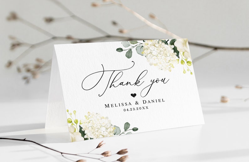 May include: White thank you card with a floral design featuring white hydrangeas and green leaves. The text "Thank you" is written in cursive with a small heart and the names "Melissa & Daniel" and the date "04.23.20XX" below.