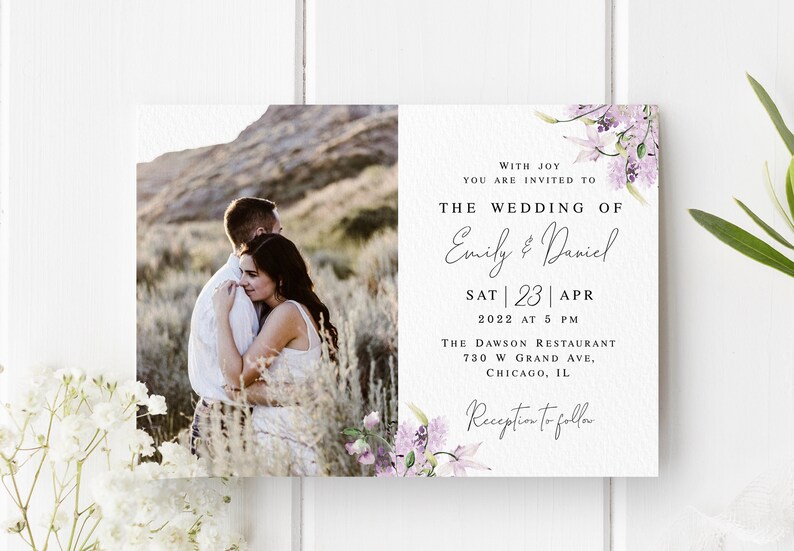 May include: Wedding invitation with a photo of a couple embracing in a field. The invitation features a floral design with purple flowers and the text "With joy you are invited to the wedding of Emily & Daniel Sat 23 Apr 2022 at 5 PM The Dawson Restaurant 730 W Grand Ave. Chicago, IL Reception to follow."