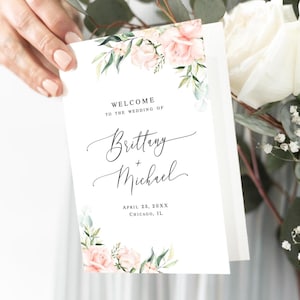 May include: A white wedding welcome sign with floral accents. The sign reads "Welcome to the Wedding of Brittany + Michael" with the date and location below. Pink roses and green leaves decorate the corners.