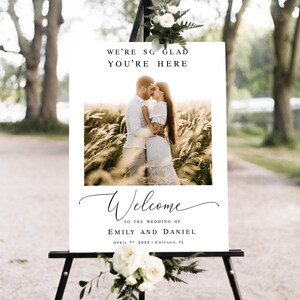 May include: A wedding welcome sign with a photo of a couple in a field. The sign reads "We're so glad you're here" and "Welcome to the wedding of Emily and Daniel, April 9th, 2022, Chicago, IL."