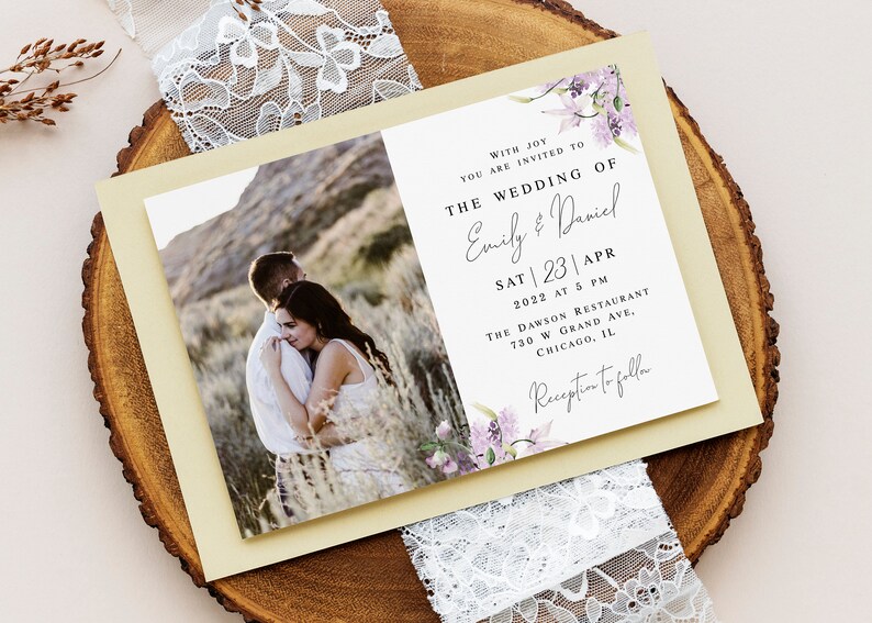 May include: Wedding invitation with a photo of a couple embracing in a field. The invitation is decorated with purple flowers and has the text "With joy you are invited to the wedding of Emily & Daniel Sat | 23 | Apr 2022 at 5 PM The Dawson Restaurant 730 W Grand Ave. Chicago, IL Reception to follow."