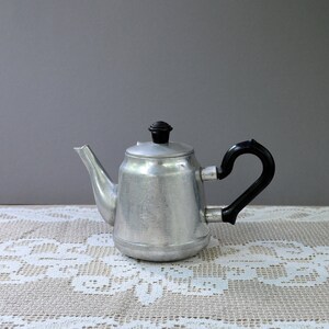 Puede incluir: Una tetera vintage de aluminio con asa y tapa negras. La tetera tiene un diseño clásico con un pico y un asa curvada. Se encuentra sobre un mantel de encaje blanco sobre un fondo gris.