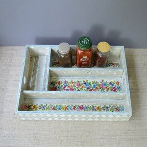 Puede incluir: Un especiero decorativo de madera con múltiples compartimentos. El estante está pintado de color crema con un estampado floral y ribete azul. Tres frascos de vidrio con especias están dentro.