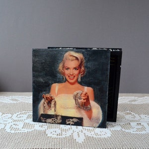 Könnte beinhalten: Ein quadratisches Kunstwerk im Vintage-Stil mit einem Porträt von Marilyn Monroe in einem weißen Kleid und Schmuck. Das Kunstwerk ist auf einem schwarzen Holzblock montiert, mit dunklem Hintergrund. Das Bild steht auf einer weißen Spitzentischdecke.