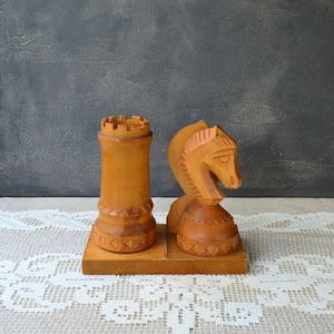 May include: Wooden chess set featuring a rook and a knight. The pieces are carved from light-colored wood and rest on a shared base. The rook has a crenellated top, and the knight is sculpted with a horse's head.