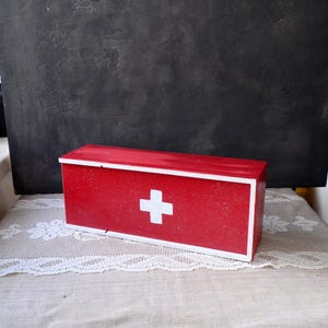 May include: A red wooden first aid box with a white cross emblem on the front. The box has a rectangular shape with white trim and a lid. The box is sitting on a white lace doily.